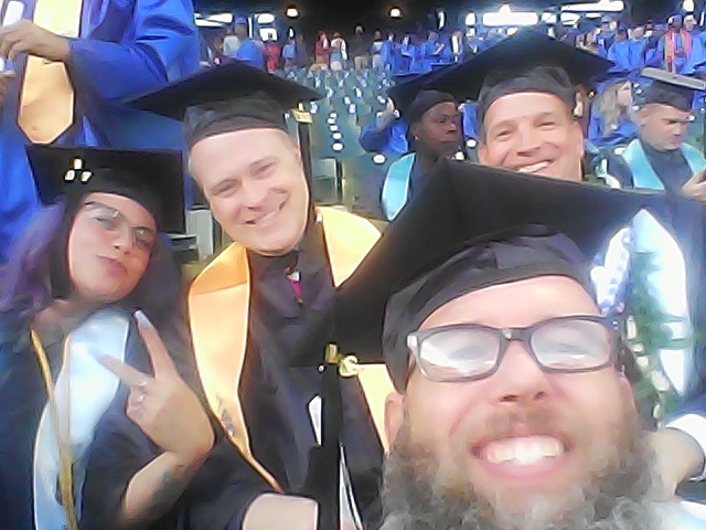 A selfie of me at commencement, wearing a cap and gown.  Behind me are a few of my classmates.