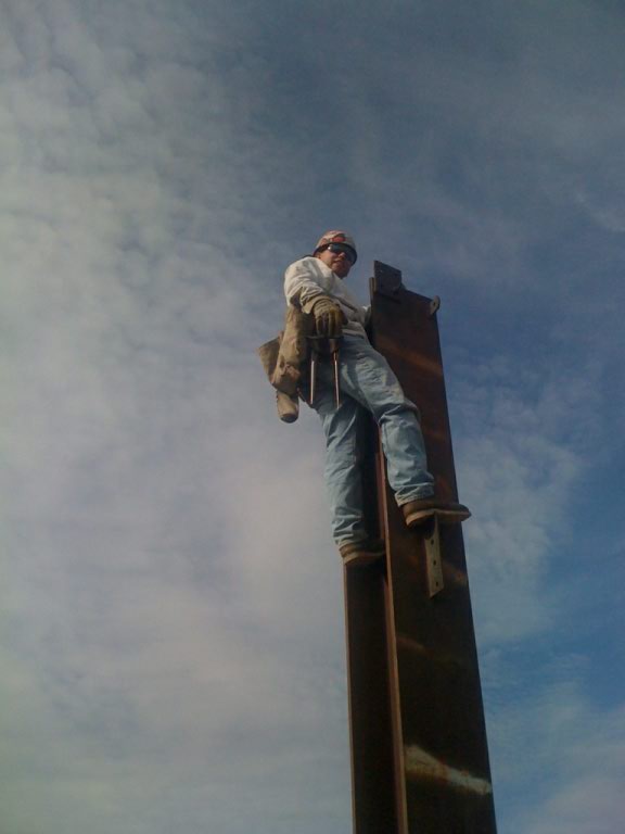 Coolest welding teacher in the three galaxies. A man is at the top of a vertical beam wearing a tool belt. the background is blue sky.