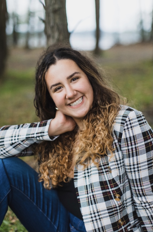 Picture of a woman with curly brown hair with highlights wearing business-casual clothes while sitting in park.