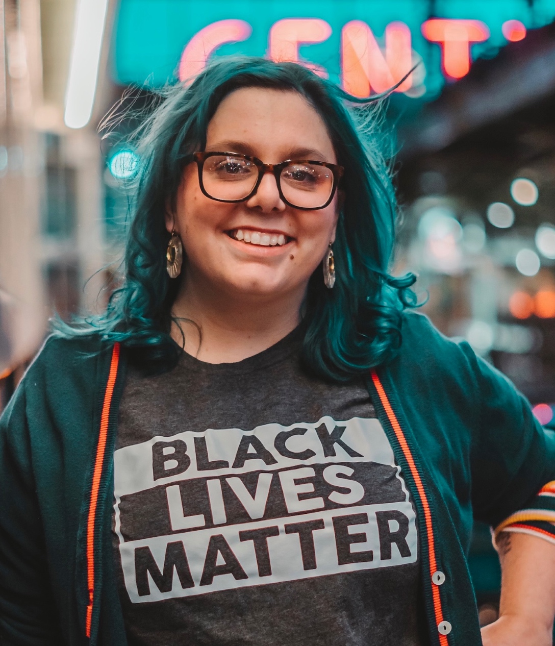 Person with blue hair and Black Lives Matter shirt standing against city background