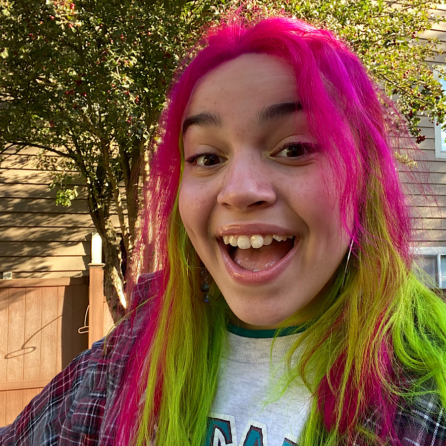 Gracie has pink and green hair, and is wearing a red and gray flannel shirt.