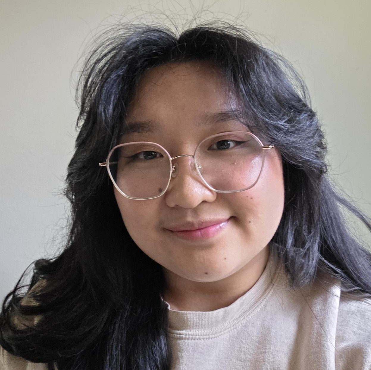 A picture of Gracie smiling at the camera. She has long, black hair and is wearing a cream-colored sweater and thin, rose gold glasses.